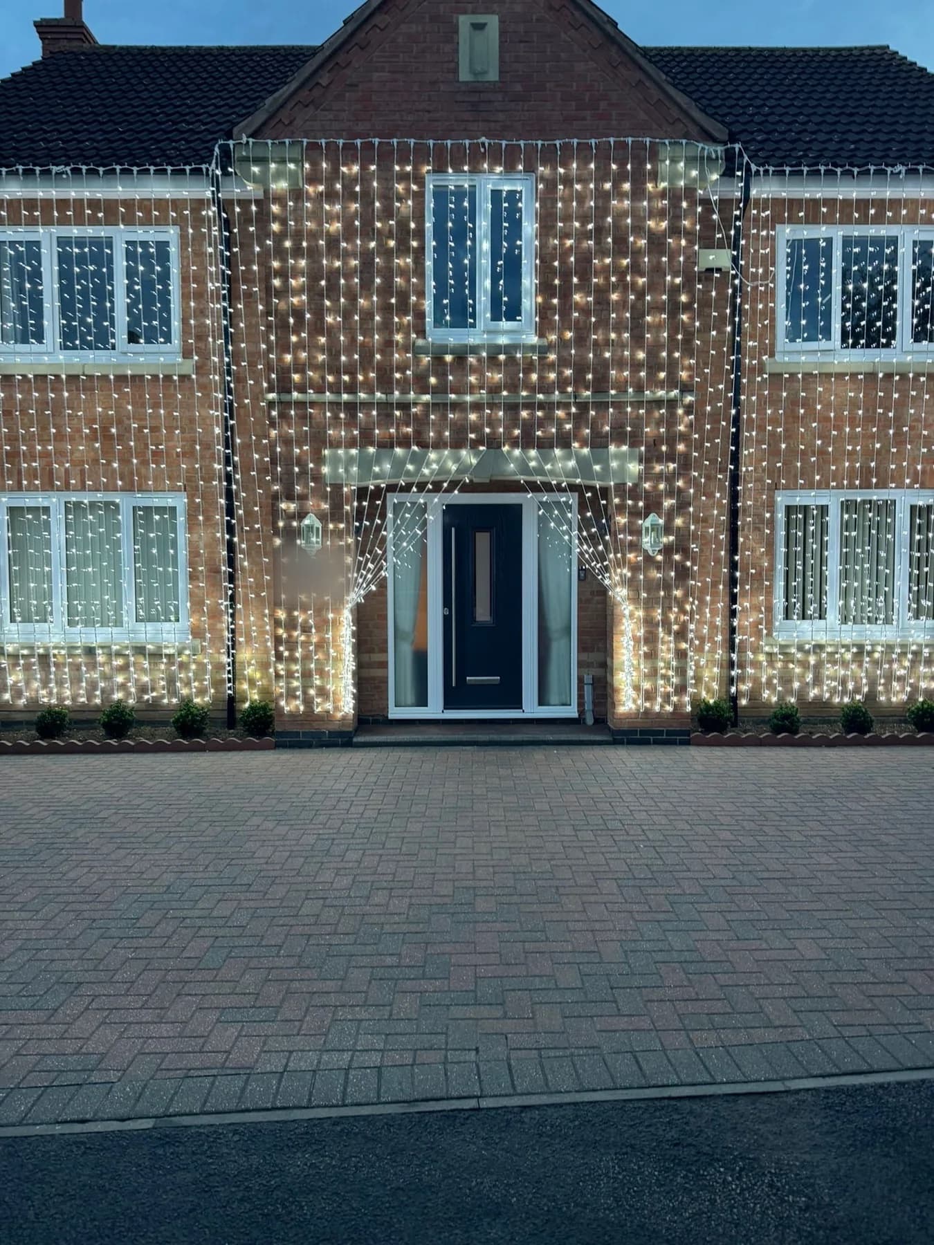 Brick house façade at dusk dressed in warm-white fairy-light curtains, with lights draped around the front door