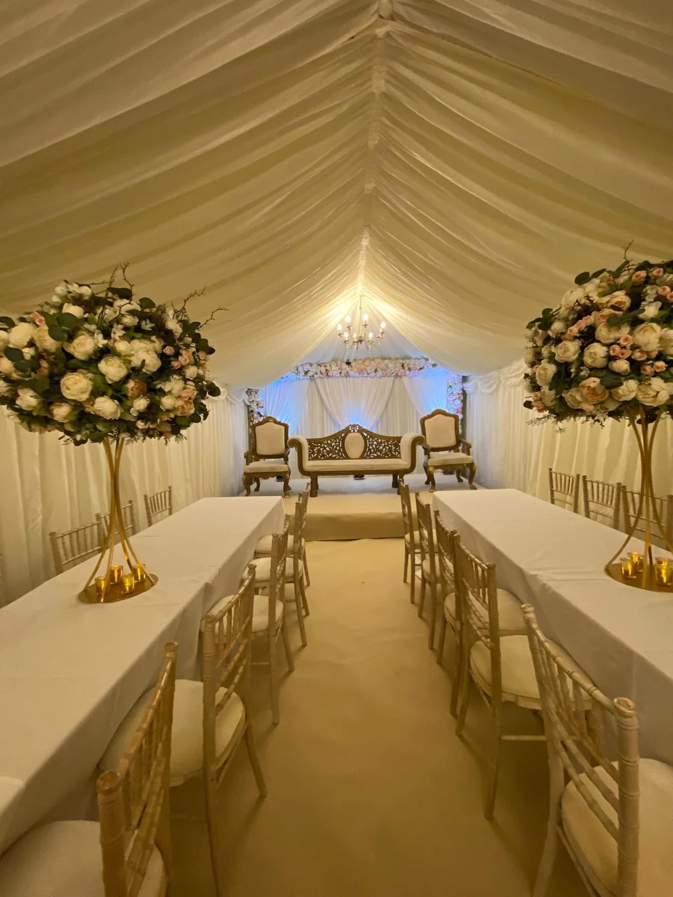 Ivory draped marquee with a chandelier, long tables dressed in white, limewash chairs and tall gold floral centrepieces leading to a raised stage