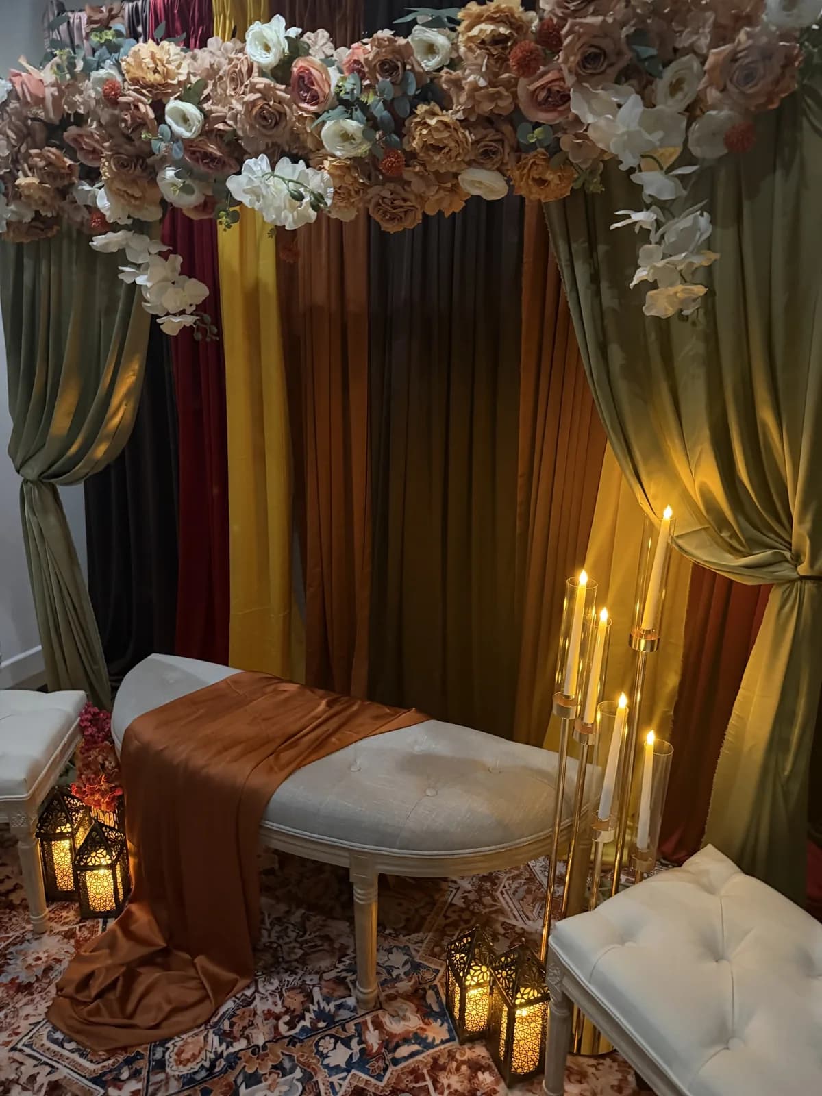 Candlelit mehndi stage with layered velvet drapes in rust, mustard and sage, a peach and ivory rose arch, a cream bench with a rust runner and Moroccan lanterns