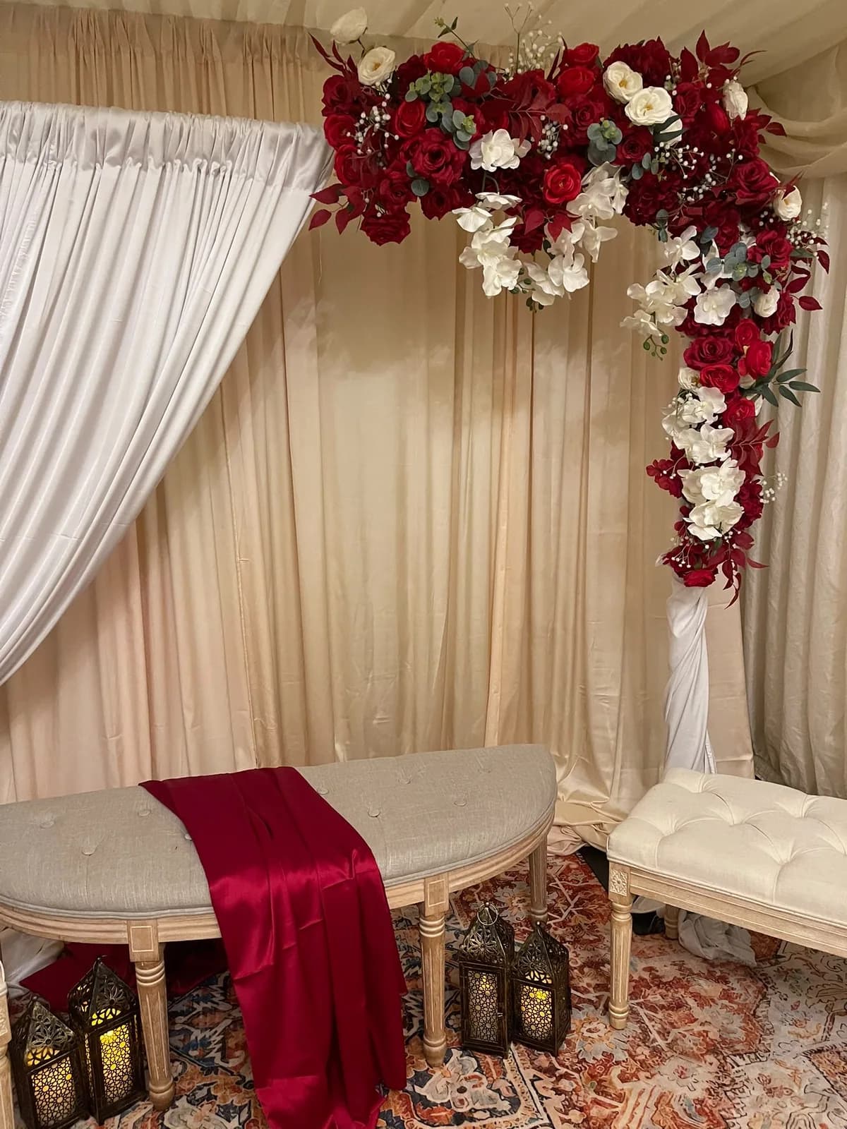 Baraat stage with ivory drapes, a red rose and white orchid floral arch, a linen bench with a deep red satin runner and Moroccan lanterns