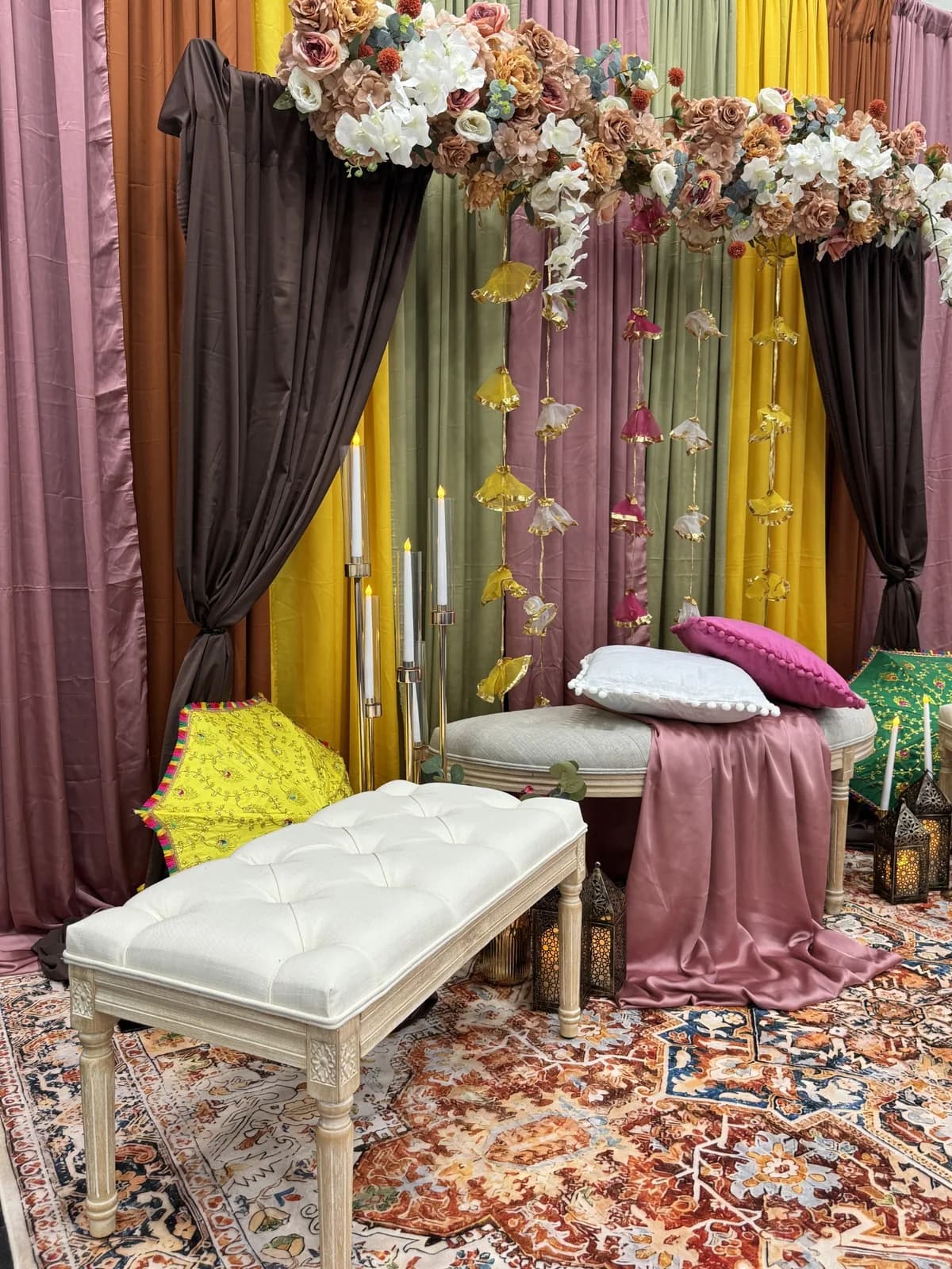Side view of a mehndi stage with a cream tufted bench, dusky pink satin runner, pink and white cushions, pillar candles, lanterns and an embroidered parasol