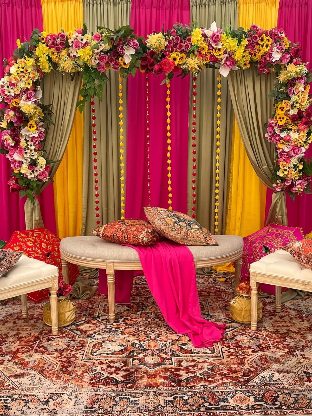 Complete mehndi stage with hot pink, yellow and sage drapes, a full pink and yellow floral arch, pom-pom garlands, a linen bench with paisley cushions and embroidered parasols